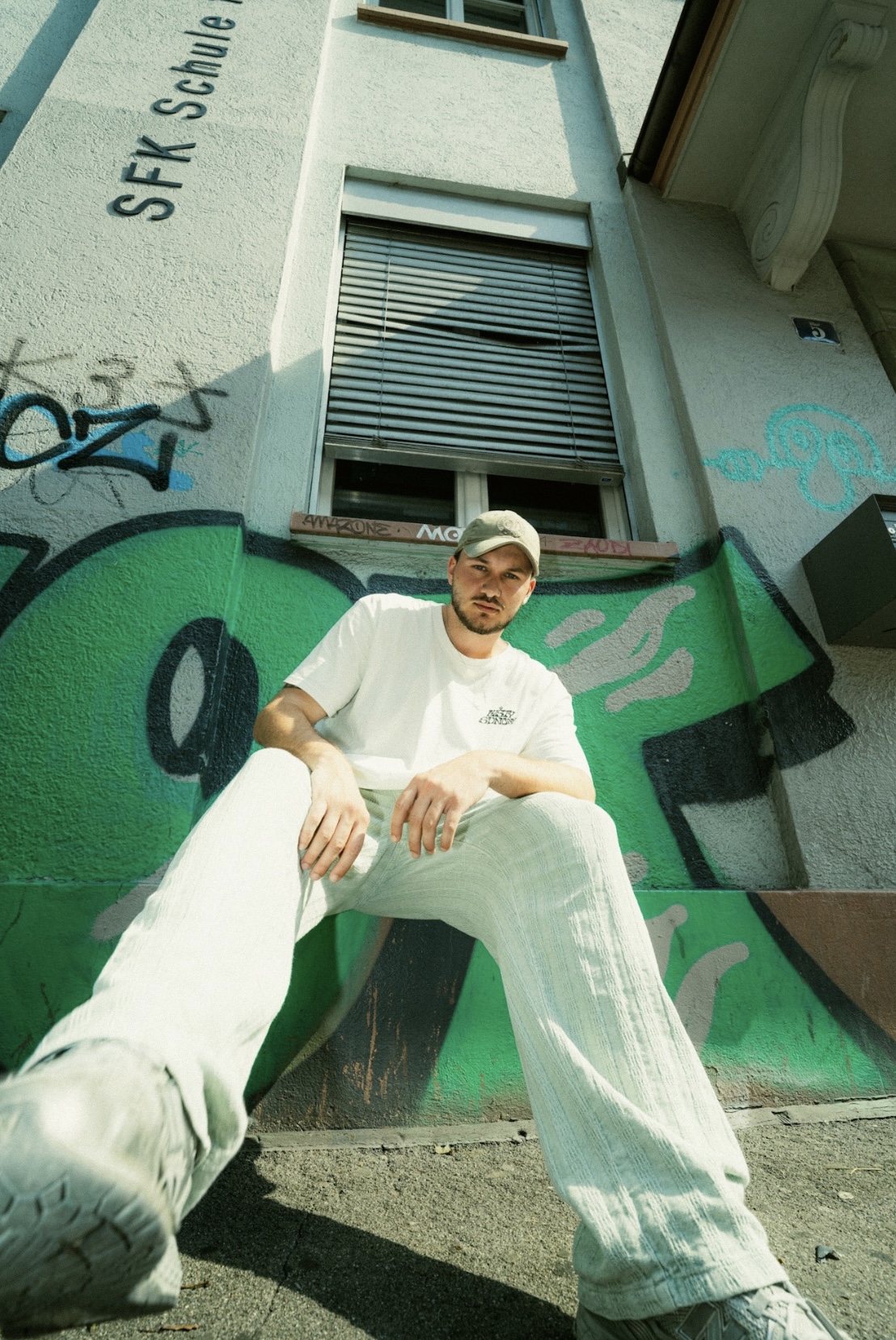 :DARREN portrait — white tee, cap, sitting in front of a graffiti wall